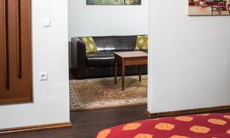 A modern bedroom with a bed and a red blanket. Through an open passage, a cozy living area with a sofa and a table is visible.