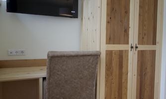 A cozy room with a desk, a chair, and a wooden wardrobe. A television hangs on the wall.