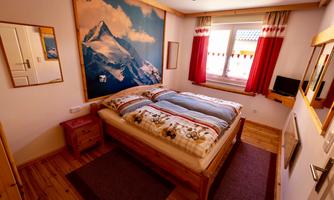 A cozy bedroom with a large bed and a panoramic picture of a mountain on the wall. The windows are adorned with red curtains and the wooden floor gives a warm atmosphere.