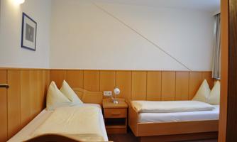 A simple room with two single beds and a bedside lamp. The walls are wood-paneled and the room is bright and friendly.