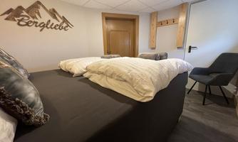A cozy room with a large bed and a wooden-decorated wall featuring the inscription "Bergliebe". There is a chair and a door in the background that leads to another room.