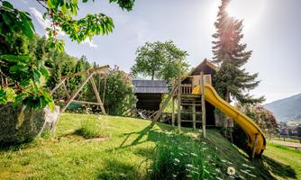 A playground with a slide and a swing in a green garden. In the background, there is a playhouse and the sun is shining.