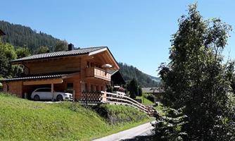 A pretty wooden house in the mountains, surrounded by green meadows. The road gently runs by, and in the background, more mountains can be seen.