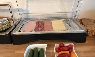 A buffet with various types of sausages and cheeses in a container. Next to it is a bowl of fresh vegetables and cherry tomatoes.
