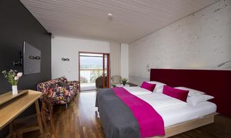 A modern bedroom with a large bed and colorful pillows. The room features large windows overlooking the outside and stylish furniture.