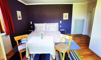 A cozy bedroom with a double bed, a table, and two chairs. The walls are painted purple and there is a beautiful wooden floor.