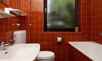A small bathroom with red tiles and a window. There is a bathtub, a sink, and a toilet.