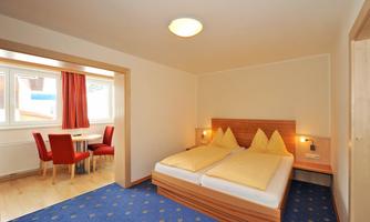 A cozy bedroom with a double bed and two pillows. In the background, there is a dining area with red chairs and a table.
