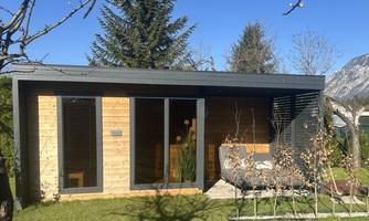 A modern wooden house with large windows and a clear blue sky. The surrounding landscape is green and inviting.