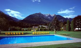 A beautiful swimming pool with yellow loungers on a green meadow. In the background, majestic mountains and blue sky are visible.