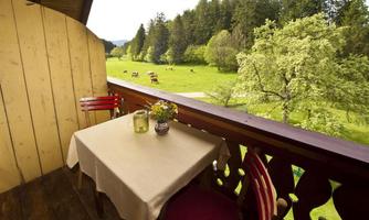 A cozy balcony with a table and chairs overlooks a green meadow with cows. In the background, there are trees and a picturesque mountain landscape.