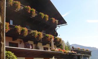 A charming wooden house with balconies decorated with blooming plants. In the foreground, there is a car and the surroundings are mountainous and sunny.