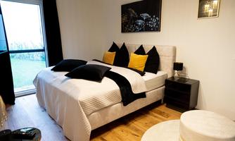 A stylish bedroom with a large bed and colorful pillows. The room is minimally furnished and has a modern view of the garden.