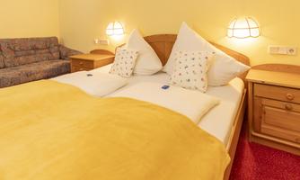 A cozy bedroom with a large bed and yellow blankets. In the background, there is a couch and warm light creates an inviting atmosphere.