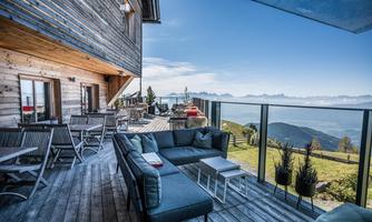 A modern terrace with a cozy seating area and a view of the mountains. The sunny sky and the green spaces in the background create a relaxed atmosphere.