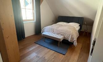 A cozy bedroom with wooden floors and sloping ceilings. The room is bright and features a double bed and a window with curtains.