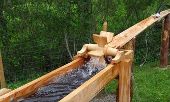 A wooden water trough directs flowing water through a green landscape. The mechanism is handmade and displays rustic charm.