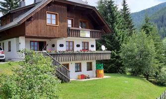 A cozy wooden house surrounded by trees and meadows. In the foreground, a few cars and colorful flower boxes can be seen.