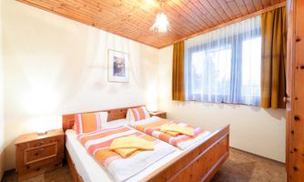 A cozy bedroom with a double bed and warm wooden furniture. The large windows let in a lot of light, and the room feels inviting.