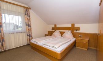 A simple bedroom with a double bed and two pillows. The room has a window with curtains and wooden furniture.