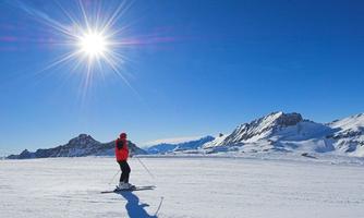 A skier stands on a wide, snow-covered area under a radiant blue sky. The sun is shining brightly, and the mountains are visible in the background.