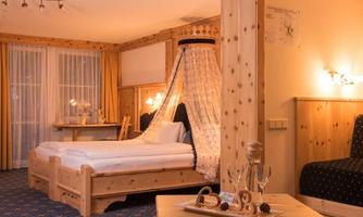 A cozy bedroom with wooden furniture and a four-poster bed. The room is bright and cheerfully decorated, with large windows and a modern table.