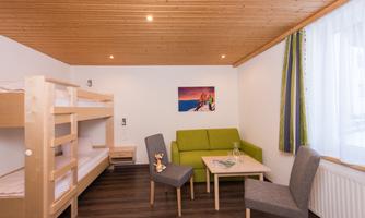 A cozy room with a bunk bed, a green sofa, and a small table. Bright walls and a wooden ceiling create a friendly atmosphere.