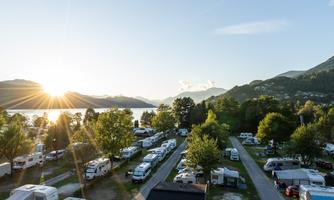A campsite by the lake with many RVs and a beautiful sunset in the background. The surroundings are green with trees and gentle hills.