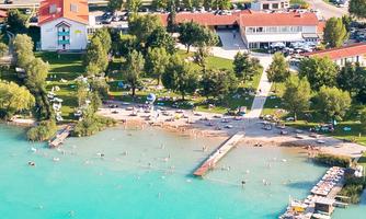 A clear, blue lake with a beach and a dock. Surrounded by green trees and some buildings.