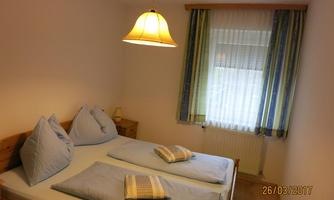 A simple bedroom with a double bed and two blue pillows. The room has a window with curtains and pleasant lighting.