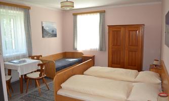 A bright room with a double bed and a single bed. The room features a table with chairs and natural light sources through large windows.