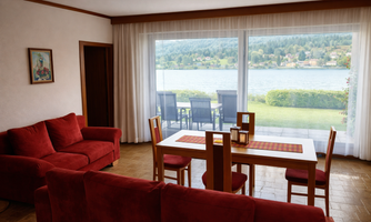 A cozy living room with red sofas and a table. Through large windows, one looks out at a lake and a green landscape.