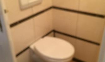 A small bathroom with a toilet and a trash can. The walls are tiled and have a simple design.