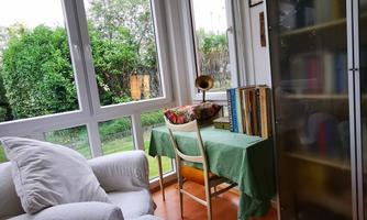 A cozy room with a large window front and a view of greenery. A table with a desk lamp and a bookshelf complete the decor.