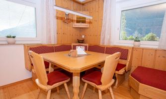 A cozy dining area with a wooden table and red upholstered chairs. The walls and window frames are made of light wood, and there is plenty of natural light.