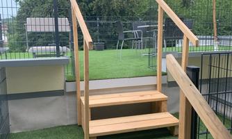 Stairs made of wood lead to a green area with artificial grass. In the background, some garden furniture and a parasol can be seen.