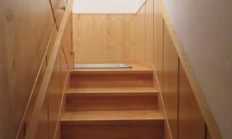 A bright wooden staircase leads upstairs. Pictures hang on the wall, and the room feels cozy.