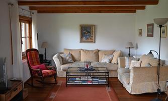 A cozy living room with a beige couch and a red armchair. A coffee table stands in the center, surrounded by window light and wooden details.