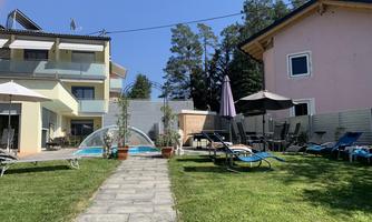 A beautiful garden with a pool, sun loungers, and shaded parasols. In the background, two houses can be seen, surrounded by trees.