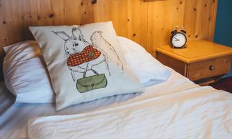 A cozy bedroom with a wooden bed frame and a pillow featuring an animal motif. Next to the bed is a nightstand with a clock and a lamp.