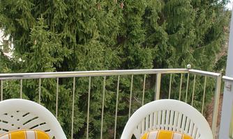 A small balcony with a round table and two white chairs that have orange-striped cushions. In the background, tall green trees are visible.