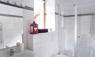 A modern bathroom with white tiles and a shower. A red lantern sits on the windowsill and purple towels are ready.