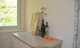 A bright kitchen area with a wooden board, several bottles of oil, and a vase with flowers. Orange scatter decor is on the countertop.
