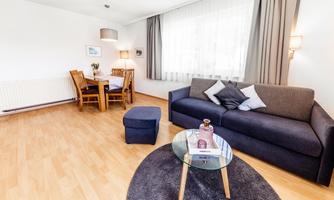 A cozy living room with a gray sofa and a round table. There is a dining area with wooden furniture and plenty of daylight from the windows.