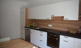 A modern kitchen with white cabinets and a wooden cladding. It includes a sink, a stove, and a refrigerator, as well as a wooden dining table.