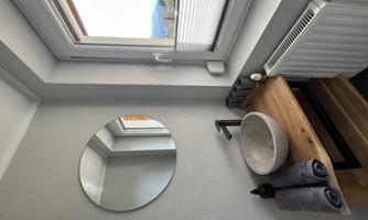 A modern bathroom with a round mirror and a simple stone sink. On the wooden shelf, there are towels and a radiator is visible.