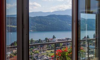 View from a window overlooking a picturesque lake and the surrounding mountains. Colorful flowers adorn the balcony.