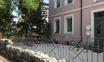 A pretty house with a pink facade and white windows. In front of the house, there is a small garden with a metal fence and a stone wall.
