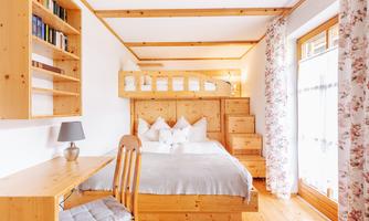 A cozy room with wooden furniture and a double bed as well as a high bed. Large windows and a bright decor create an inviting atmosphere.