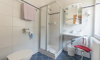 A modern bathroom with a shower, sink, and toilet. The walls are light tiled, and there are towels and a sink chair.